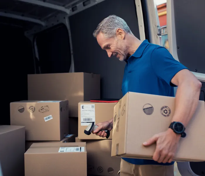 A man scans boxes with a barcode reader inside a delivery van, wearing a blue polo shirt and a watch.