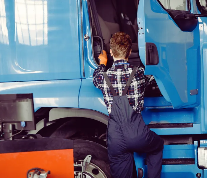 Person in overalls climbs into a blue truck cabin inside a garage.