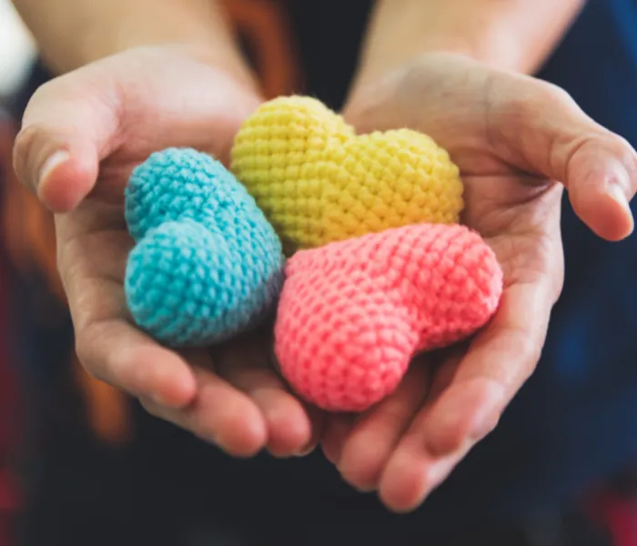 Hands holding three knitted heart shapes in blue, yellow, and pink.