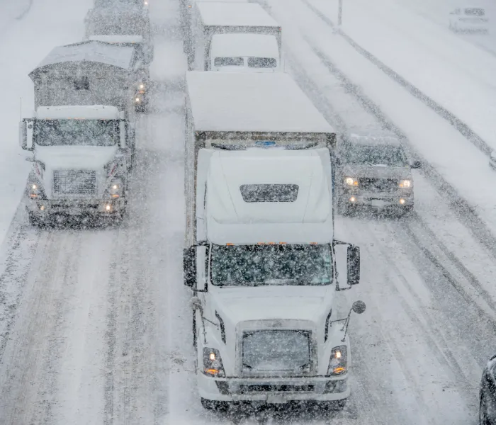 Heavy snow falls on a highway, where multiple trucks and cars are driving slowly in reduced visibility conditions.