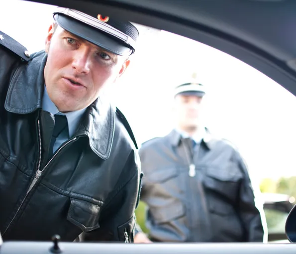 Two police officers in black uniforms are looking inside a car, one close to the window, the other standing in the background.