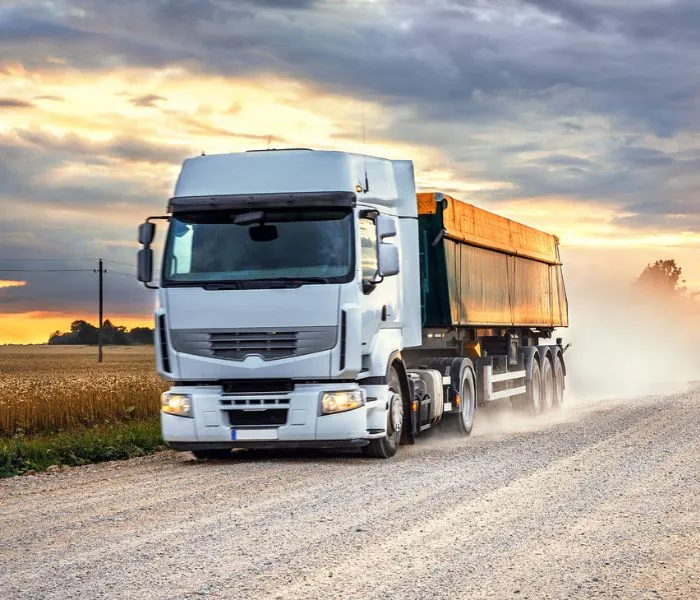 A white semi-truck with a trailer drives on a gravel road through a rural landscape at sunset.